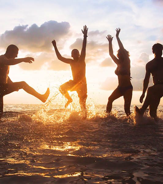 group of happy friends having fun together on the beach at sunset, jumping and dancing with water splash in the sea, silhouettes of people